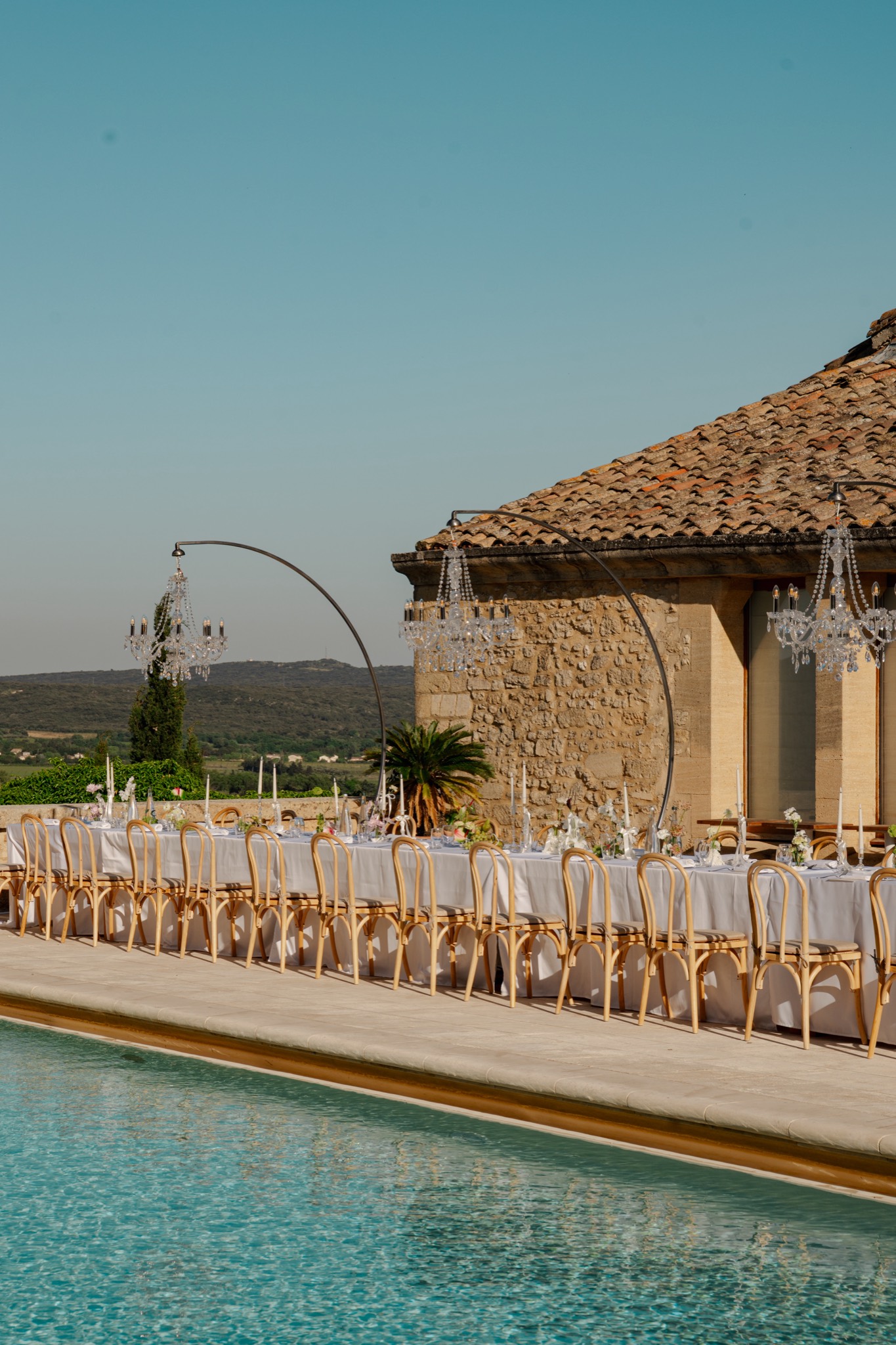 A long dinner table set beside a pool at a stone villa, lit for the evening